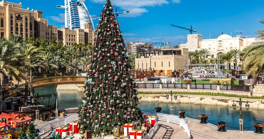 christmas lighting dubai - a large christmas tree with presents in front of a city, christmas tree and christmas lighting in front of burj al arab, christmas tree and lighting in Souq Madinat
