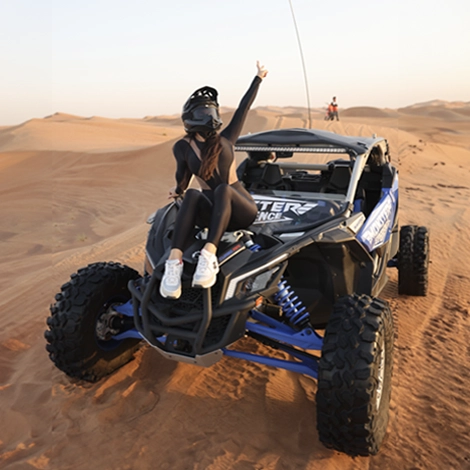 A girl wearing helmet and sitting on top of black and blue buggy with posing with peace sign in the desert